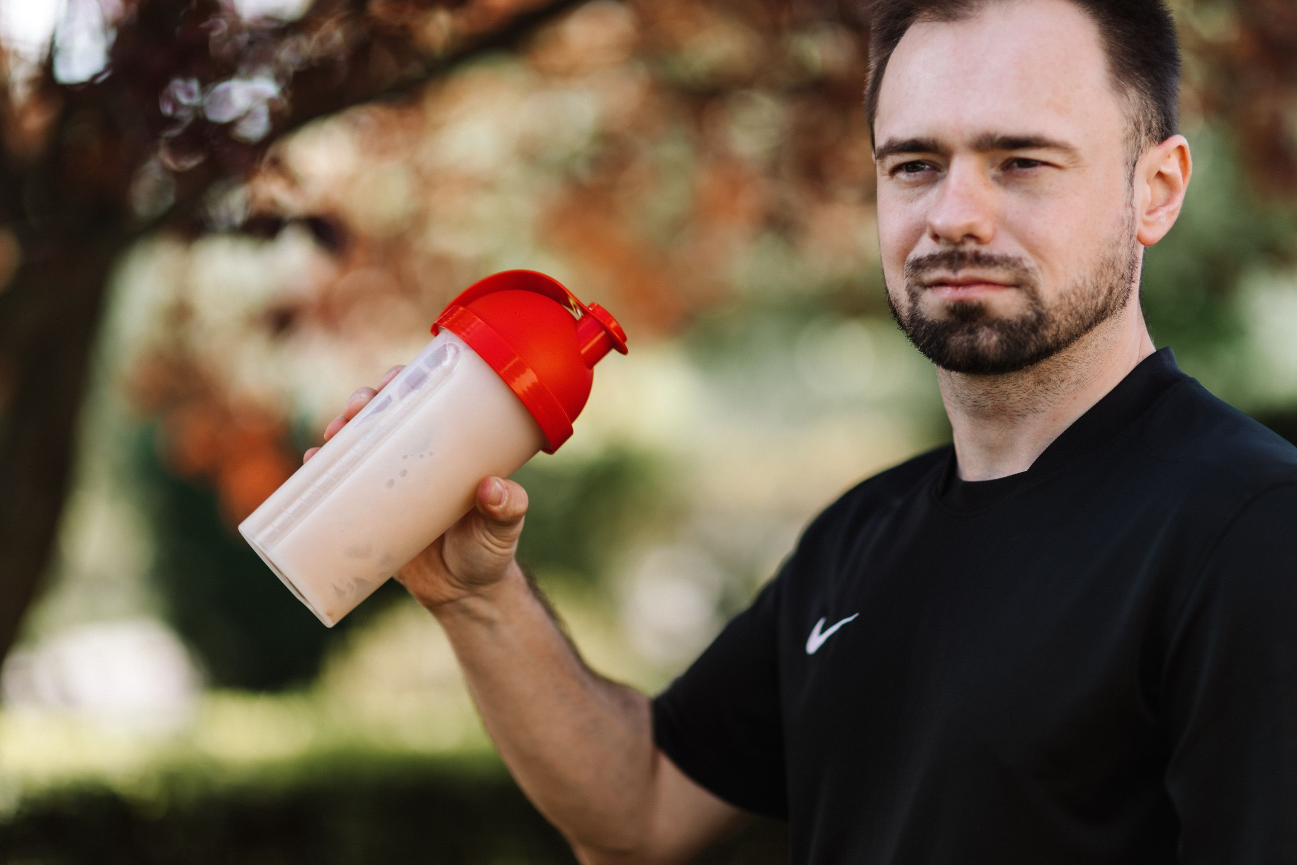 What Role Does Protein Play in Our Body Adult man holds a protein shake in a red lid tumbler outdoors, wearing a black shirt.