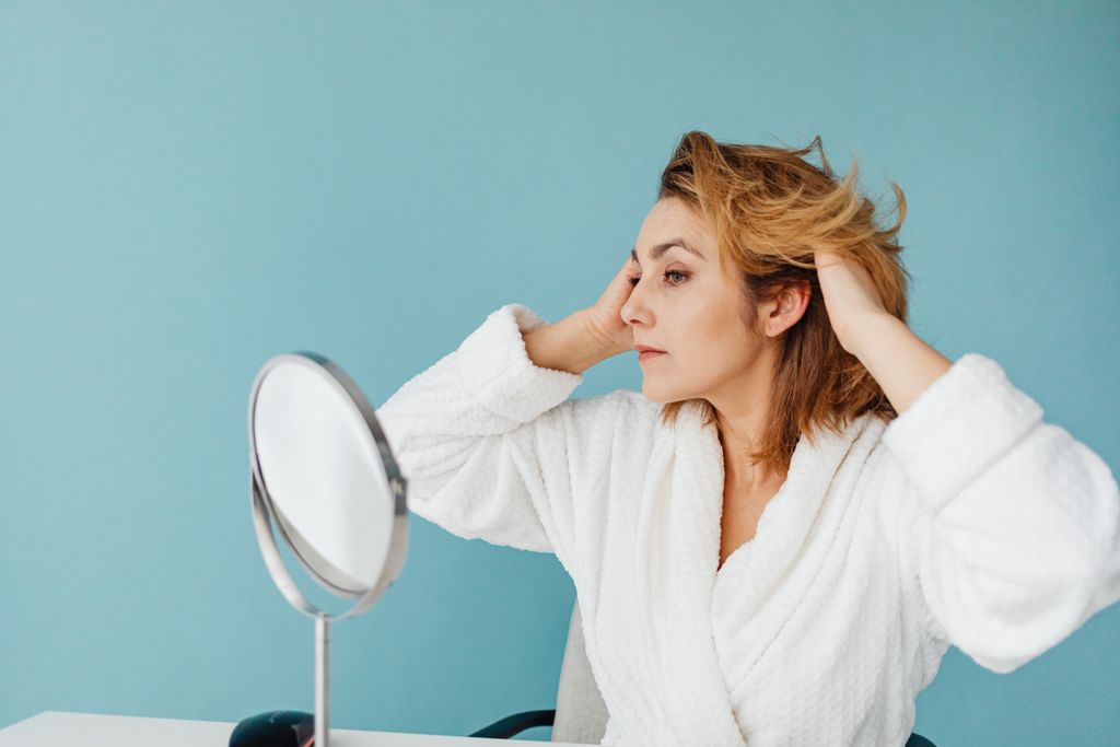 Adult woman in a bathrobe fixing her hair in front of a mirror against a blue background.