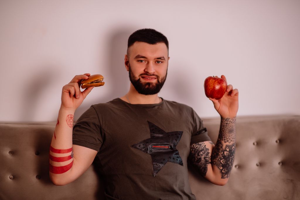 Bearded man pondering diet choice with apple and burger. Indoors with tattoos visible.