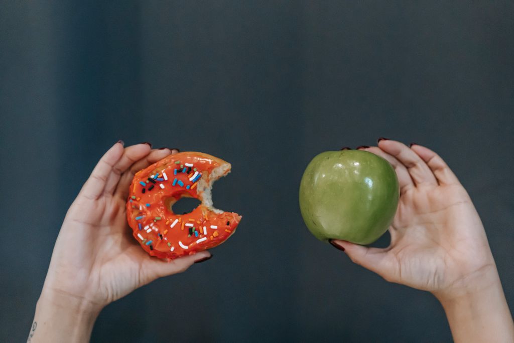 Crop unrecognizable young female demonstrating bitten high calorie doughnut and healthy green apple against gray background