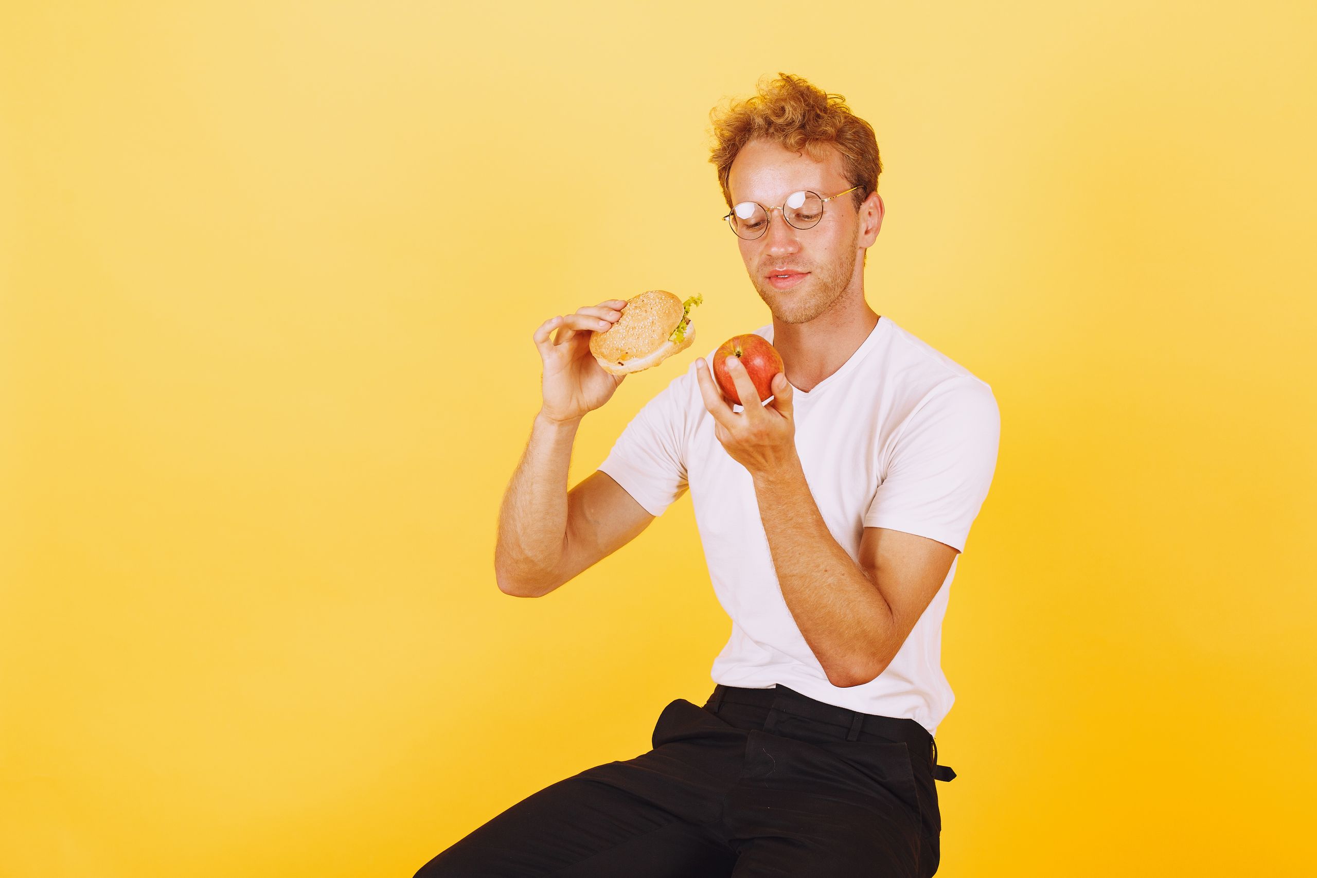 Man in white shirt holding a burger and apple on yellow background.